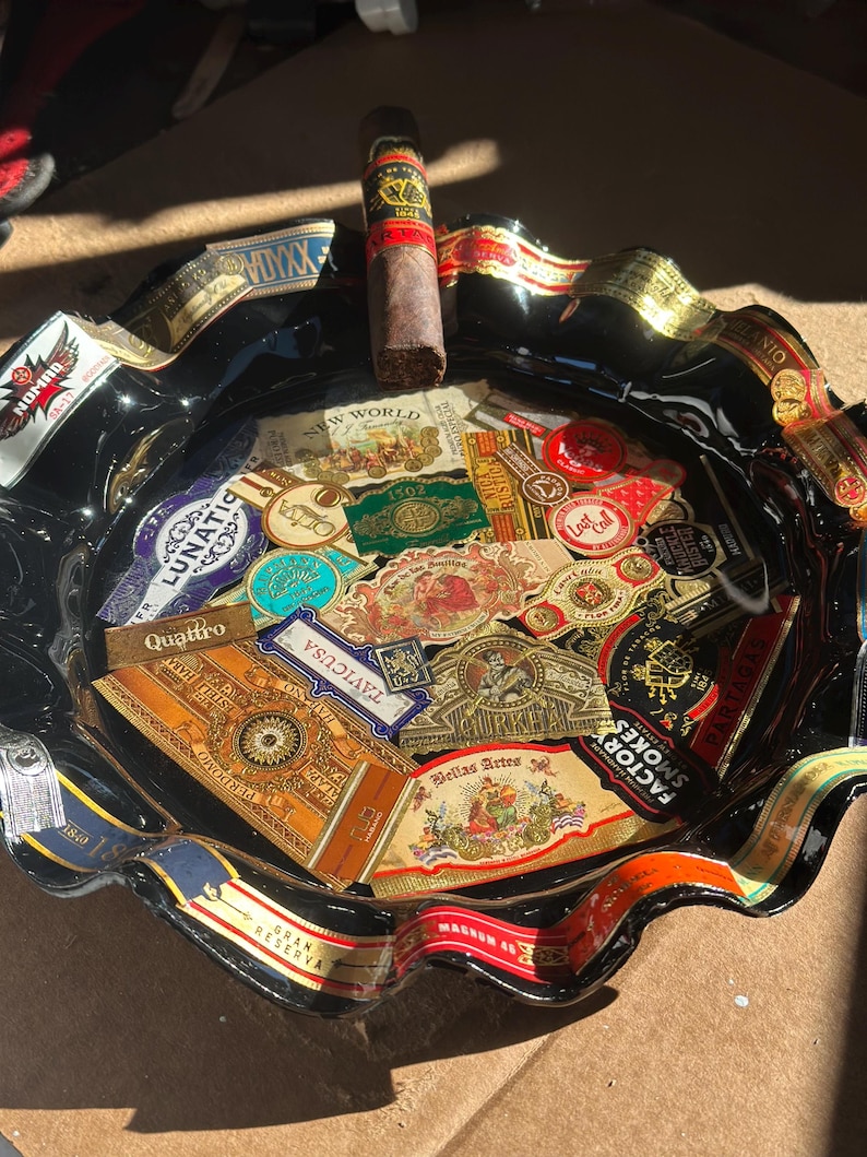 K&ouml;nnte beinhalten: Ein schwarzer Aschenbecher aus Glas mit gewelltem Rand, der eine Collage aus verschiedenen bunten Zigarrenringen auf der Oberfl&auml;che zeigt. Eine einzelne braune Zigarre mit einem rot-goldenen Ring liegt auf dem oberen Rand des Aschenbechers, der zahlreiche Markennamen und komplexe grafische Designs aufweist.