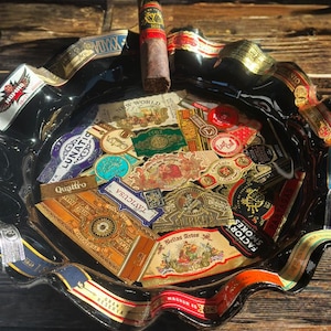 K&ouml;nnte beinhalten: Ein schwarzer Aschenbecher aus Glas mit gewelltem, geschwungenem Rand, verziert mit verschiedenen bunten Zigarrenringen, die im Glas eingebettet sind. Eine einzelne braune Zigarre mit schwarz-goldener Banderole liegt auf dem oberen Rand des Aschenbechers, der mit einer Collage aus zahlreichen Zigarrenetiketten im Vintage-Stil gef&uuml;llt ist.
