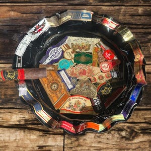 K&ouml;nnte beinhalten: Ein runder Aschenbecher aus schwarzem Glas mit gewelltem Rand, der mit verschiedenen bunten Zigarrenringen verziert ist. Das Innere des Aschenbechers ist mit einer Collage aus zahlreichen Zigarrenetiketten in verschiedenen Formen und Farben gef&uuml;llt, wobei eine braune Zigarre auf dem Rand liegt.