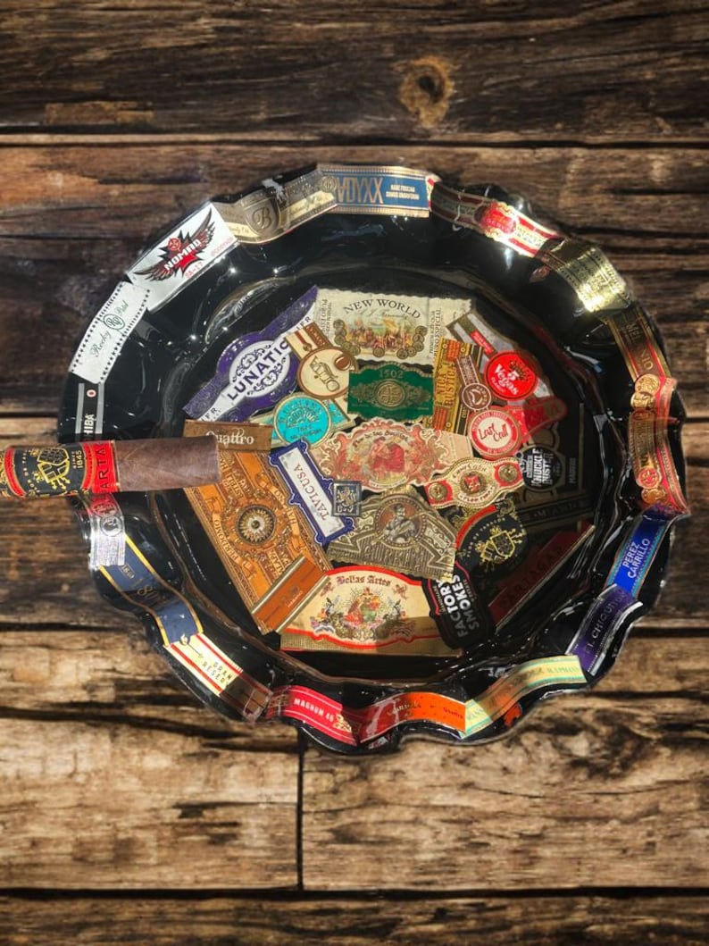 K&ouml;nnte beinhalten: Ein runder Aschenbecher aus schwarzem Glas mit gewelltem Rand, der mit verschiedenen bunten Zigarrenringen verziert ist. Das Innere des Aschenbechers ist mit einer Collage aus zahlreichen Zigarrenetiketten in verschiedenen Formen und Farben gef&uuml;llt, wobei eine braune Zigarre auf dem Rand liegt.