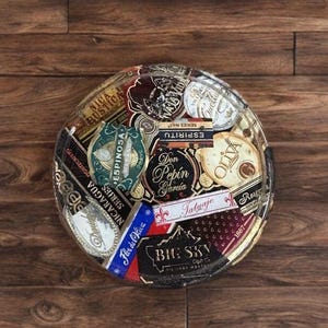 Puede incluir: Un posavasos redondo y decorativo con un collage de etiquetas de cigarros. Las etiquetas son de varios colores, incluyendo rojo, azul, dorado y blanco, y presentan texto y gr&aacute;ficos. El posavasos est&aacute; sobre un fondo de madera.