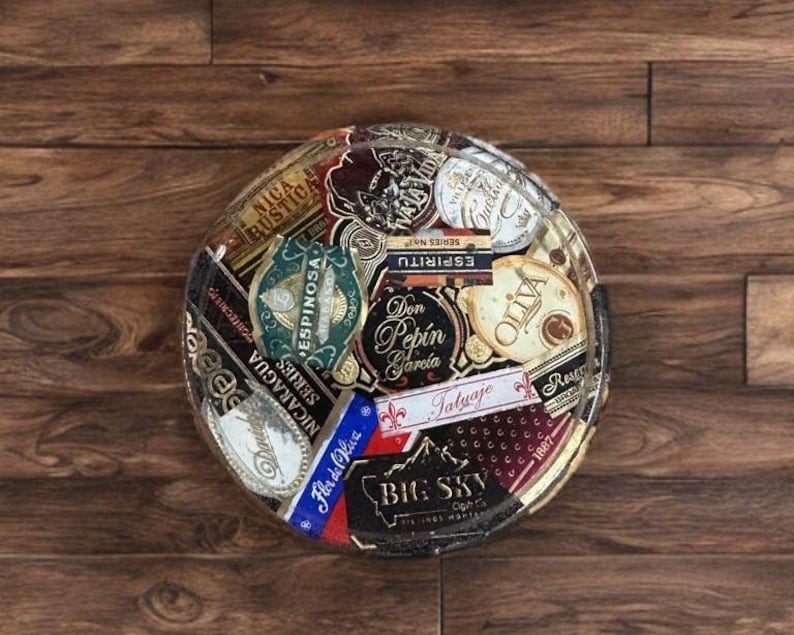 Puede incluir: Un posavasos redondo y decorativo con un collage de etiquetas de cigarros. Las etiquetas son de varios colores, incluyendo rojo, azul, dorado y blanco, y presentan texto y gr&aacute;ficos. El posavasos est&aacute; sobre un fondo de madera.