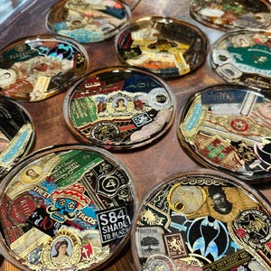 Puede incluir: Una colecci&oacute;n de posavasos redondos de resina transparente. Cada posavasos presenta un collage de coloridas etiquetas y logotipos de cigarros, con detalles dorados. Los posavasos est&aacute;n dispuestos sobre una superficie de madera, mostrando una variedad de dise&ntilde;os y marcas.