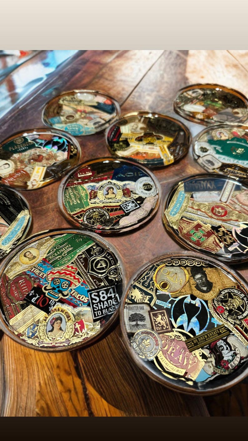 Puede incluir: Una colecci&oacute;n de posavasos redondos de resina transparente. Cada posavasos presenta un collage de coloridas etiquetas y logotipos de cigarros, con detalles dorados. Los posavasos est&aacute;n dispuestos sobre una superficie de madera, mostrando una variedad de dise&ntilde;os y marcas.