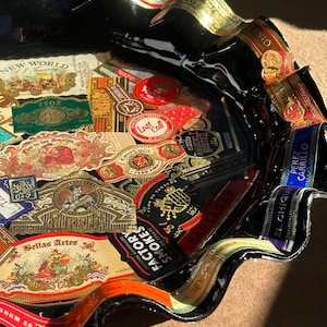 K&ouml;nnte beinhalten: Ein schwarzes Glasschalen-Tablett mit gewelltem Rand, das mit einer Collage aus verschiedenen bunten Zigarrenringen verziert ist. Die Oberfl&auml;che ist mit zahlreichen Papieretiketten bedeckt, darunter Marken wie Gurkha, Partagas, New World und Bellas Artes.