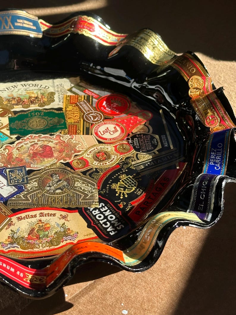 K&ouml;nnte beinhalten: Ein schwarzes Glasschalen-Tablett mit gewelltem Rand, das mit einer Collage aus verschiedenen bunten Zigarrenringen verziert ist. Die Oberfl&auml;che ist mit zahlreichen Papieretiketten bedeckt, darunter Marken wie Gurkha, Partagas, New World und Bellas Artes.
