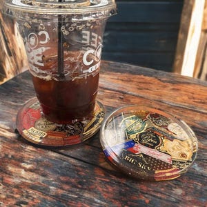 Puede incluir: Una taza de pl&aacute;stico transparente con caf&eacute; helado descansa sobre un posavasos. El posavasos y un segundo son redondos, con una parte superior de resina transparente y un collage de etiquetas coloridas. Los posavasos est&aacute;n sobre una superficie de madera.