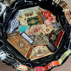 K&ouml;nnte beinhalten: Ein rundes, schwarzes Tablett mit gewelltem Rand, das eine Collage aus verschiedenen bunten Zigarrenringen zeigt. Die Ringe weisen diverse Markennamen und Logos auf, darunter New World, Vegas, Lunatic, Gurkha, Partagas und Perdomo, die auf der Fl&auml;che angeordnet sind.