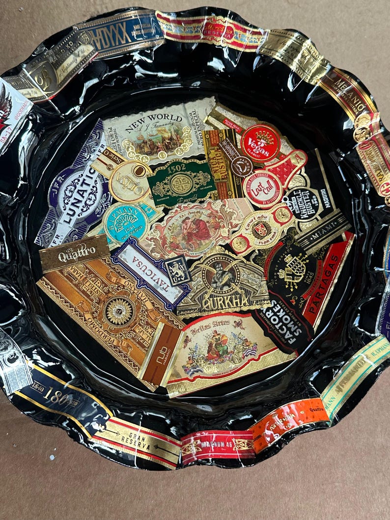 K&ouml;nnte beinhalten: Ein rundes, schwarzes Tablett mit gewelltem Rand, das eine Collage aus verschiedenen bunten Zigarrenringen zeigt. Die Ringe weisen diverse Markennamen und Logos auf, darunter New World, Vegas, Lunatic, Gurkha, Partagas und Perdomo, die auf der Fl&auml;che angeordnet sind.