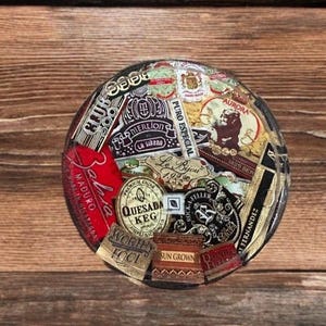 Puede incluir: Un posavasos redondo con un collage de coloridas etiquetas de cigarros. Las etiquetas incluyen textos como "Aurora", "Quesada Keg" y "Maduro". El posavasos est&aacute; sobre un fondo de madera.