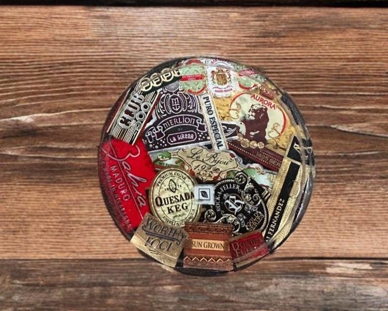 Puede incluir: Un posavasos redondo con un collage de coloridas etiquetas de cigarros. Las etiquetas incluyen textos como "Aurora", "Quesada Keg" y "Maduro". El posavasos est&aacute; sobre un fondo de madera.