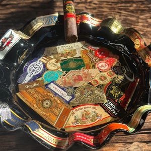K&ouml;nnte beinhalten: Ein schwarzer Aschenbecher aus Glas mit gewelltem Rand, verziert mit einer Collage aus verschiedenen bunten Zigarrenringen. Eine einzelne braune Zigarre mit schwarz-goldener Banderole liegt auf dem oberen Rand des Aschenbechers.