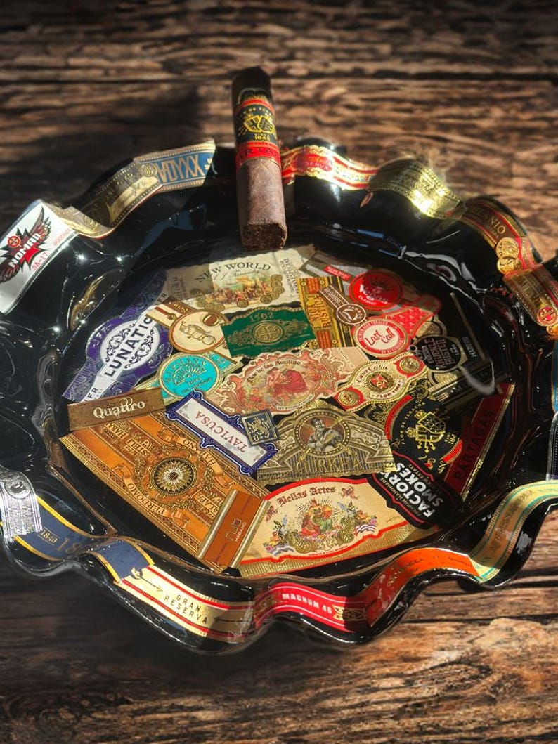 K&ouml;nnte beinhalten: Ein schwarzer Aschenbecher aus Glas mit gewelltem Rand, verziert mit einer Collage aus verschiedenen bunten Zigarrenringen. Eine einzelne braune Zigarre mit schwarz-goldener Banderole liegt auf dem oberen Rand des Aschenbechers.