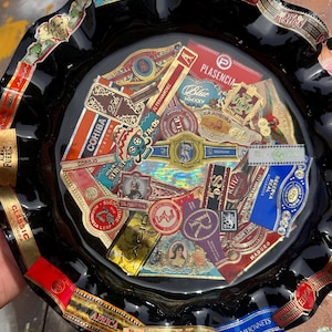 K&ouml;nnte beinhalten: Ein runder Aschenbecher aus schwarzem Glas mit gewelltem Rand, der eine Collage aus verschiedenen bunten Zigarrenringen unter einer gl&auml;nzenden Oberfl&auml;che zeigt. Die Ringe zeigen diverse Markennamen und Logos wie Cohiba, Plasencia und La Aurora.