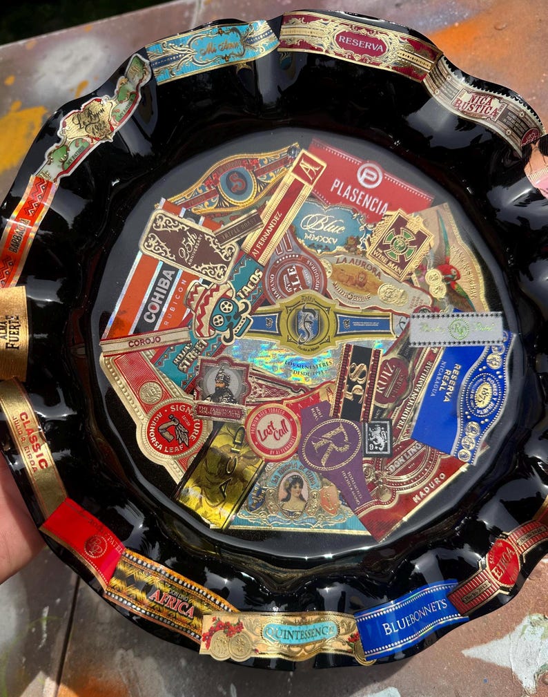 K&ouml;nnte beinhalten: Ein runder Aschenbecher aus schwarzem Glas mit gewelltem Rand, der eine Collage aus verschiedenen bunten Zigarrenringen unter einer gl&auml;nzenden Oberfl&auml;che zeigt. Die Ringe zeigen diverse Markennamen und Logos wie Cohiba, Plasencia und La Aurora.