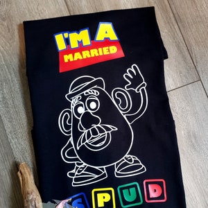 May include: Black t-shirt featuring a white outline of Mr. Potato Head and the text "I'm a Married Spud" in colourful block letters.