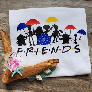 May include: White t-shirt featuring a silhouette of Toy Story characters holding umbrellas, with the word "FRIENDS" spelled out below.