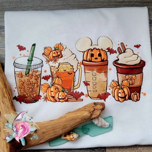 May include: White t-shirt featuring a graphic of four autumn-themed drinks with pumpkin spice and Mickey Mouse ears. The drinks are decorated with pumpkins and bats.
