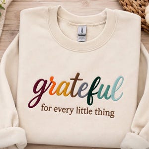 May include: A cream-colored crewneck sweatshirt featuring the embroidered word grateful in a multi-colored gradient of maroon, orange, yellow, brown, dark green, and light blue, with the text for every little thing in brown below it.