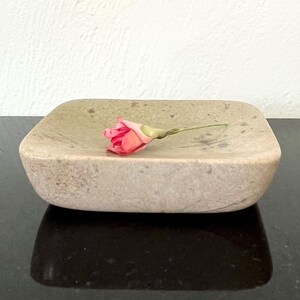 May include: A square, beige stone soap dish with rounded edges. A pink rosebud rests on the dish. The dish is made of a speckled stone material and sits on a dark surface.