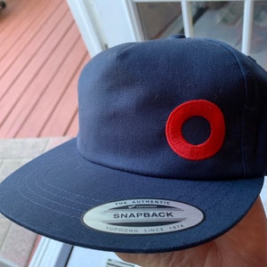May include: A navy blue baseball cap with a red circle logo. The logo has a smaller circle inside. The cap has a white sticker on the back that says "The Authentic Yupoong Snapback Yupoong Since 1974".