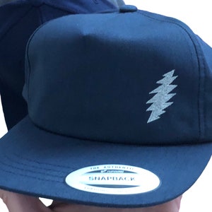 May include: Navy blue snapback hat with a silver lightning bolt design on the front. The hat has a flat brim and a white tag that reads "The Authentic Snapback". The hat is a classic style.