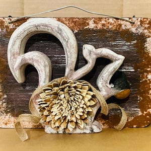 Puede incluir: Un colgante de pared rústico de madera con una escultura estilizada del número "62" en blanco, con un girasol decorativo y una cinta de arpillera. El fondo es marrón y blanco desgastado. Se adjunta un alambre de metal para colgar.