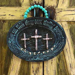 Könnte beinhalten: Ovale Holzschild mit drei Kreuzen und dem Text "Gib uns heute unser tägliches Brot". Das Schild ist dunkelblau mit türkisfarbenen Akzenten und einem türkisfarbenen Perlen-Griff. Der Hintergrund ist eine verwitterte gelb-braune Holztür.