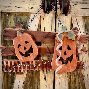 Puede incluir: Un letrero rústico de Halloween de madera con dos calabazas naranjas talladas con caras sonrientes. Las calabazas están unidas a una valla de madera marrón con una vid blanca decorativa y una pequeña valla de estacas. El letrero cuelga de una cadena.