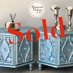 May include: Two matching blue painted side tables with ornate carved details and crystal knobs. Each table has a top surface with decorative items, including a diffuser and floral arrangements. The word "SOLD" is prominently displayed.