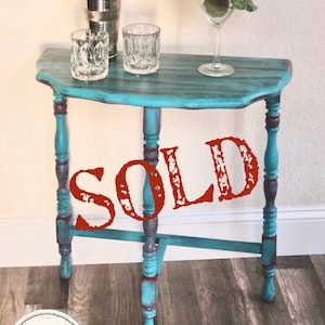 May include: A teal and brown painted side table with a half-moon top. The table has turned legs and a distressed finish. Two patterned glasses, a cocktail shaker, and a martini glass with flowers sit on top. The word "SOLD" is printed on the table.