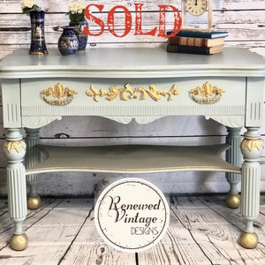 May include: A light blue vintage console table with gold accents. The table features ornate gold drawer pulls and decorative trim. The legs are fluted and have gold ball feet. The word "SOLD" is in red across the top. A clock and vases are on the table.