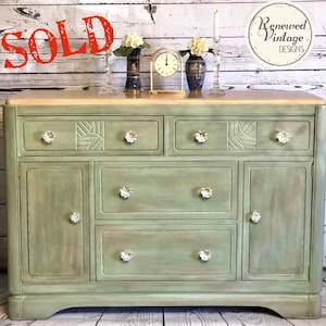 May include: A vintage-style, sage green dresser with crystal knobs. The dresser has two small drawers, two doors, and three larger drawers. The top is a light wood color. The word "SOLD" is stamped in red across the top left corner.