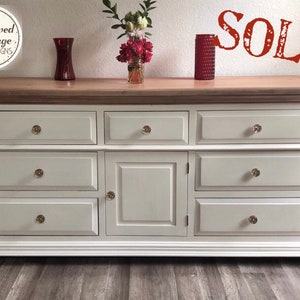 May include: A white dresser with seven drawers and a central cabinet. The top is a light wood color, and the drawers have crystal knobs. The word "SOLD" is stamped in red on the wall above the dresser. The dresser is from "Renewed Vintage Designs".