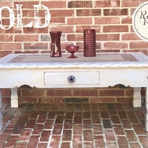 May include: A white distressed coffee table with a drawer and star-shaped hardware. The tabletop features a decorative border and a central panel. The table has a rustic, vintage aesthetic. The word "SOLD" is visible in the image.
