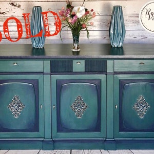 May include: A teal and navy blue painted vintage sideboard with three doors and drawers. The doors feature ornate silver-colored hardware. Two teal vases and a vase of flowers sit on top. The word "SOLD" is written across the top.