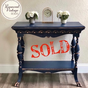 May include: Navy blue console table with gold accents. The table features a lower shelf and ornate legs. The word "SOLD" is stamped across the front. The table is decorated with white flowers and a gold clock.