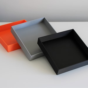 May include: Three square trays, one orange, one grey, and one black, are stacked on a white surface.