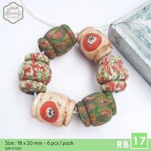 May include: A strand of six decorative beads. The beads are a mix of colors, including cream, red, and green. Some beads have a face design. The text "JAVA BEADS INDONESIA" is in the upper left corner. The text "Size: 18 x 20 mm - 6 pcs / pack" is at the bottom.
