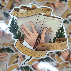 Op de afbeelding: Een witte sticker met een hand die een gesloten boek vasthoudt met een gouden lint waarop "Something is coming" staat. Het boek heeft een gouden symbool op de kaft. De hand houdt een bruin koord vast. De sticker heeft groene bladeren aan beide kanten van het boek. Het lint onder het boek zegt "You can feel it. Can't you?"