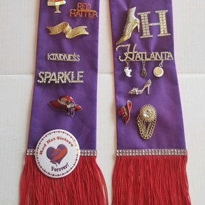 May include: Purple sash with red fringe and various rhinestone pins and charms. The pins include the words "I Love Dancing", "Magic", "50 Shades of Red", "Red Hatter", "Kindness", "Sparkle", "Virginia Crowns", "V", "M", "H", "Atlanta", and "Red Hat Sisters Forever!"