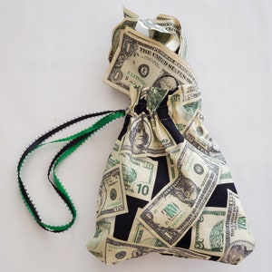 May include: A black drawstring bag with a green drawstring. The bag is made of fabric and has a pattern of dollar bills printed on it. The bag is tied with a green ribbon and has a dollar bill peeking out of the top.