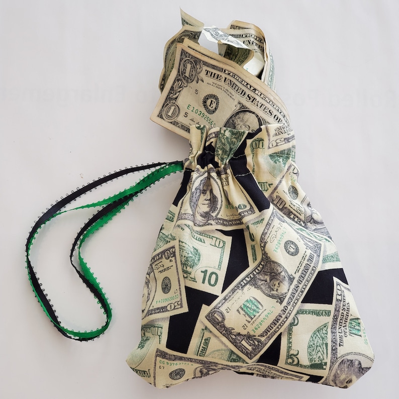 May include: A black drawstring bag with a green drawstring. The bag is made of fabric and has a pattern of dollar bills printed on it. The bag is tied with a green ribbon and has a dollar bill peeking out of the top.