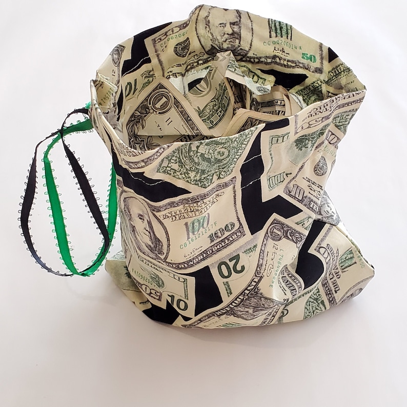 May include: A black drawstring bag with a green and black drawstring. The bag is made of fabric and has a pattern of US dollar bills.