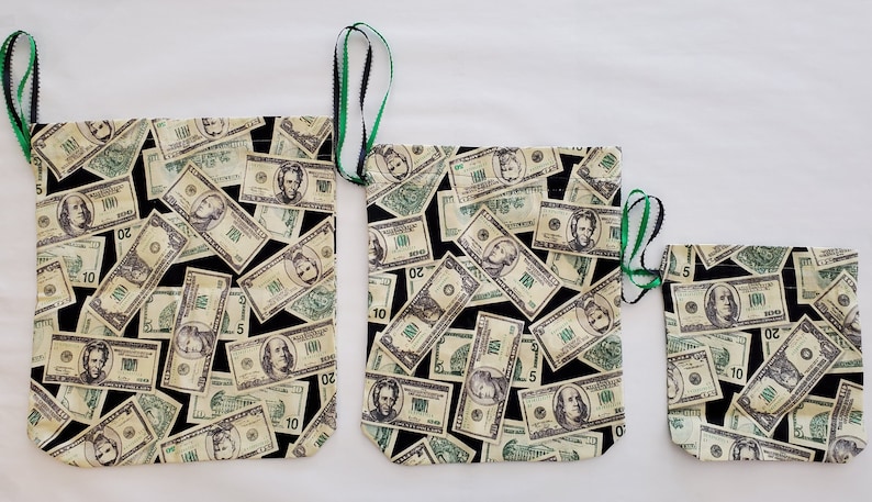 May include: Three drawstring bags with a black background and a pattern of US dollar bills in various denominations. The bags are different sizes and have green and black drawstrings.