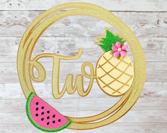 May include: Gold glitter circle cake topper with the word "Two" in gold glitter lettering. A yellow pineapple with green leaves and a pink flower is on the right side of the circle. A pink watermelon slice with black seeds is on the left side of the circle.