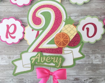 May include: A green, pink, and gold glitter cake topper with a number 2, a pineapple, an orange slice, and a strawberry. The topper has a white banner with the name Avery written in green glitter. A pink bow is attached to the bottom of the topper.