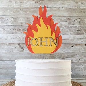 May include: A white cake with a yellow and orange paper fire topper with the name "JOHN" on it.