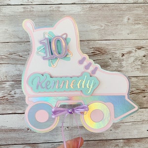 May include: A white and pink roller skate cake topper with a purple bow and a light blue and white star with the number 10 in the center. The name Kennedy is written in light blue on the skate.