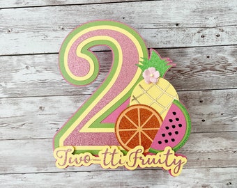 May include: A pink and yellow glitter number two cake topper with a pineapple, watermelon, and orange slice design. The topper says "Two-tti Fruity".